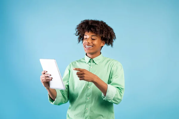 Aplicación para estudiantes. Feliz negro chico adolescente navegar por la web en la tableta PC sobre fondo azul y sonriendo —  Fotos de Stock