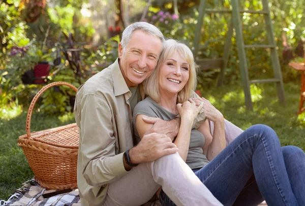 Portret pary seniorów siedzącej w ogrodzie i przytulającej się, mającej piknik na świeżym powietrzu i uśmiechniętej do kamery — Zdjęcie stockowe
