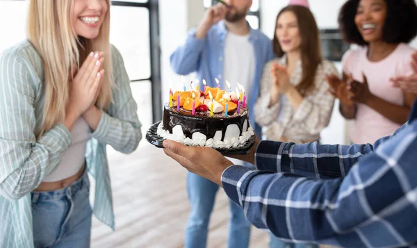 Vista de cerca de las personas alegres multirraciales que presentan pastel de cumpleaños con velas encendidas a la joven mujer rubia en casa —  Fotos de Stock