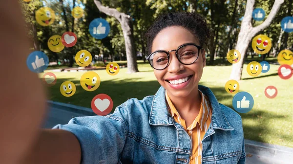 Gündelik ve gözlüklü tatmin olmuş genç siyahi kadın sosyal ağlar ve bloglar için dışarıda fotoğraf çekiyor
