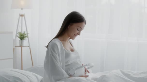 Беременная женщина держит наушники возле живота и танцует, сидя в постели — стоковое видео