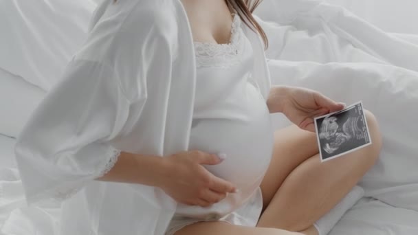 Donna incinta irriconoscibile che tiene la foto ad ultrasuoni del bambino e tocca la pancia a letto — Video Stock