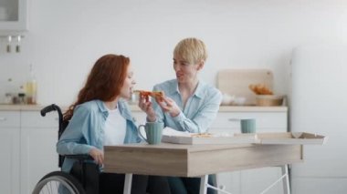 Sevgi dolu anne engelli kızını sıcak taze pizzayla besliyor. Evde tekerlekli sandalyede oturan kızıl saçlı küçük kız.