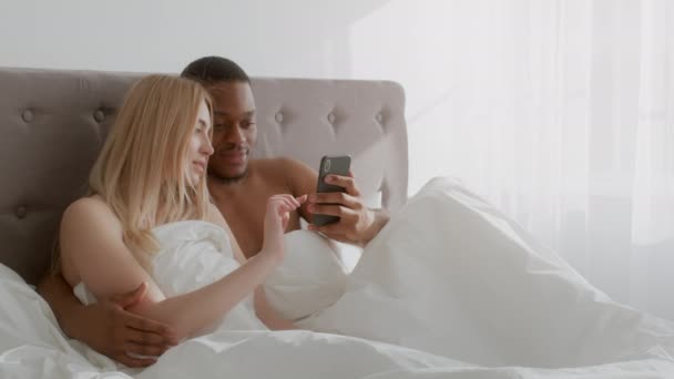 Amante casal diversificado segurando telefone celular e navegação na web nas mídias sociais, usando nova aplicação juntos, deitado na cama — Vídeo de Stock