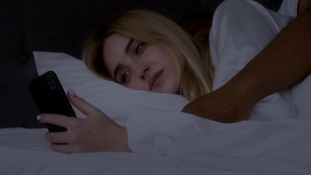 Молодая женщина болтает по смартфону в семейной постели, притворяясь спящей, когда ее черный муж просыпается ночью — стоковое видео