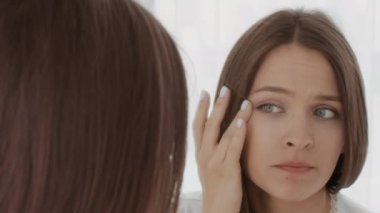 Evde aynaya bakan endişeli genç kadın, cilt durumundan memnun değil.