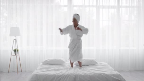 Feliz jovem afro-americano mulher vestindo roupão de banho pulando na cama em casa — Vídeo de Stock