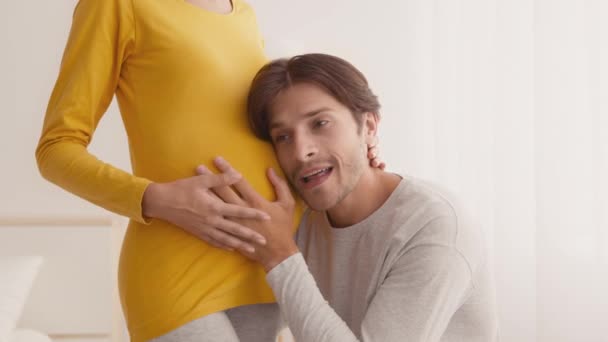 Молодой человек слушает большой живот беременной женщины, чувствуя, что ребенок ударился и улыбается жене, чувствуя себя счастливым и взволнованным — стоковое видео