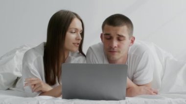 Yatakta dinlenirken dizüstü bilgisayar kullanan, online alışveriş yapan genç çift.
