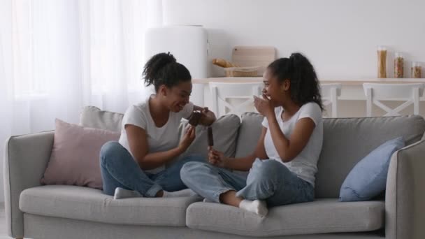 Zwei fröhliche schwarze Schwestern essen Eis und lachen zu Hause auf der Couch — Stockvideo