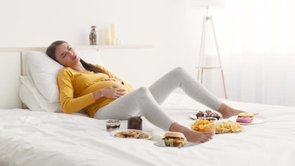 Mujer embarazada joven acostada en la cama con mucha comida, acariciando su vientre, sufriendo de acidez estomacal y estómago — Vídeo de stock