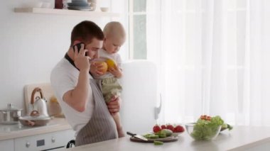 Genç baba mutfakta yemek pişirirken telefonda konuşuyor ve kucağında bebek var.