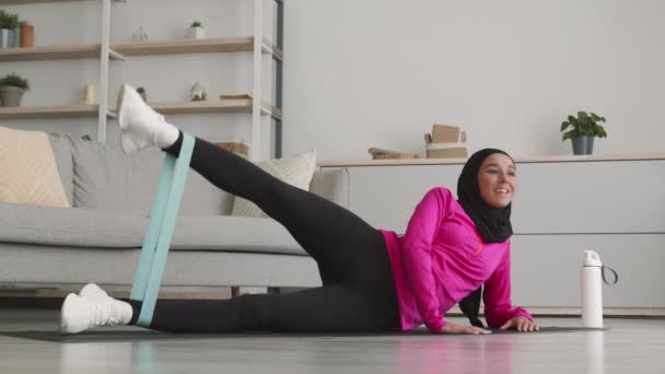 Concepto de entrenamiento corporal. Joven mujer musulmana positiva haciendo ejercicio con goma elástica, acostada en el suelo en casa — Vídeo de stock