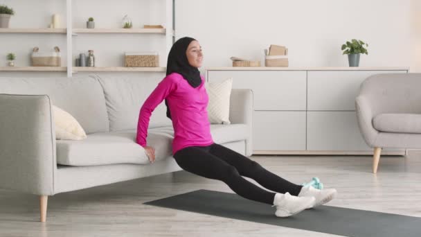 Ευτυχισμένη μεσανατολική μουσουλμάνα κυρία ακουμπά στον καναπέ και κάνει ασκήσεις push-up, γυμνάζεται στο σπίτι, ελεύθερος χώρος — Αρχείο Βίντεο