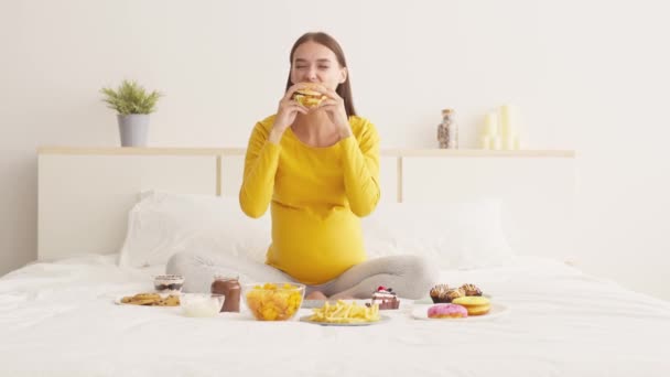 Voedseltevredenheid. Jonge gelukkige zwangere dame eten ongezonde hamburger met veel plezier, genieten van junk food delivery — Stockvideo