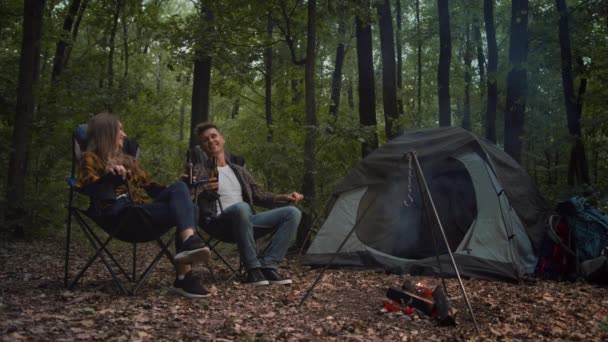 Buon weekend in campeggio. Giovane coppia seduta vicino al falò e tifo con la birra, godendo di riposo attivo nella foresta serale — Video Stock