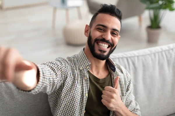Ritratto di giovane arabo felice che mostra i pollici verso l'alto gesto con entrambe le mani, seduto sul divano e sorridente a casa — Foto Stock