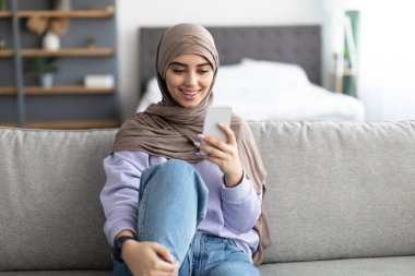 Kanepede oturan gülümseyen İslamcı kadın cep telefonu kullanıyor.