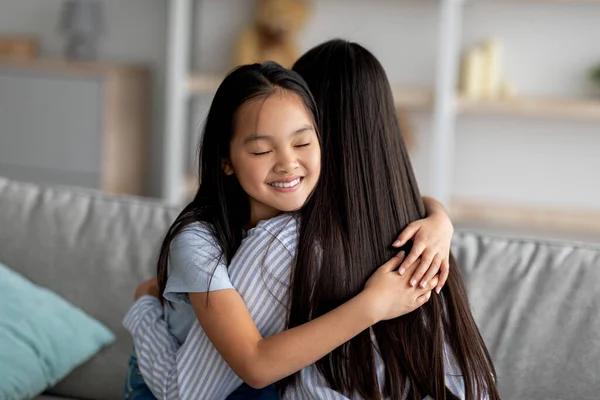 Concepto de unión. Linda chica asiática abrazando a su madre y sonriendo, sentados juntos en el sofá, primer plano retrato, espacio libre —  Fotos de Stock