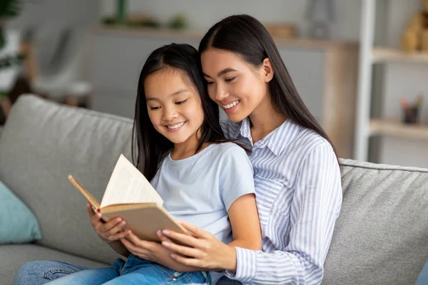 Amar joven asiática madre leer libro con su bonita hija, sentado juntos en sofá en casa — Foto de Stock