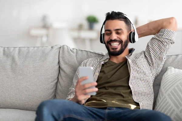 Sorrindo jovem árabe cara em fones de ouvido desfrutando de música ou áudio livro no smartphone, escolhendo playlist popular em casa — Fotografia de Stock