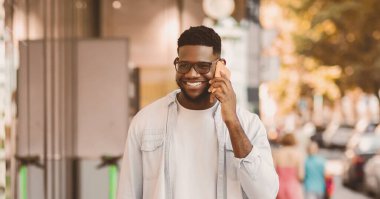 Neşeli Afro-Amerikan bir adamın tüm vücut uzunluğu cep telefonuyla konuşuyor, alışveriş torbaları tutuyor, güneşli bir günde sokakta yürüyor ve vitrinlere bakıyor. Perakende, Satış, Teknoloji, Yaşam Tarzı