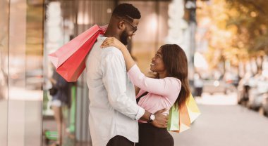 Tam vücut boy portresi, neşeli zenci kadının erkek arkadaşına sarılışının açık havada elinde alışveriş torbalarıyla duruşu. Sevgililer birbirine bakıyor, şehirde yürüyor, alışverişin tadını çıkarıyor.