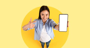 Neşeli genç bir kadın parlak sarı bir alanda duruyor, akıllı telefonunu büyük bir gülümsemeyle gösteriyor. Cihazı işaret ediyor, güven ve pozitiflik yayıyor..