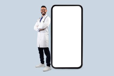 Orta Doğulu erkek doktor, beyaz üniformalı, mavi arka planda el ele tutuşan, kameraya gülümseyen. Profesyonel Tıp İşçisinin Tam Uzunluğu, Başarılı Doktor