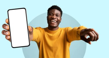 Genç bir adam elinde boş bir ekranla akıllı telefonu tutarken geniş bir gülümseme sergiliyor. Heyecanlı görünüyor, izleyiciyle ilgileniyor ve hafif bir atmosferde bir anlık mutluluğu paylaşıyor..