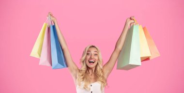 Sezonluk Satış Konsepti. Heyecanlı sarışın bir kadının portresi, ağzı açık bir şekilde bir sürü alışveriş torbası tutuyor ve kaldırıyor. Yeni aldıklarıyla mutlu, pembe stüdyo duvarında izole edilmiş.