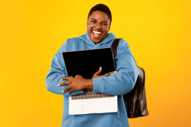 Heyecanlı Büyük-Boyutlu Siyahi Kadın Dizüstü bilgisayarı kucaklıyor Kameraya gülümsüyor Sırt çantasıyla Sarı Stüdyo Arkaplanı Üzerinde. E-Öğrenme, Çevrimiçi Teknoloji Konsepti