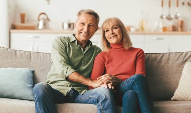 Neşeli kıdemli eşler el ele tutuşup evdeki kameraya gülümsüyor. Mutlu ve olgun bir çiftin koltukta poz vermesinin ön görüntüsü. Uzun süreli evlilik ve ilişki.