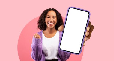 Neşeli bir kadın elinde akıllı bir telefon tutuyor, parlak bir şekilde gülümsüyor. O rahat bir kazak giyer ve pembe bir arka fonda durur, pozitiflik ve sıcaklık saçar..