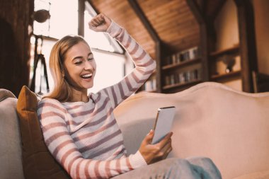 Büyük Galibiyet. Neşeli Pozitif Kadın 'ın Portresi Sevinç ve Heyecanla Titreyen Yumruk Tutan Akıllı Telefon, El Kalkıyor. Evdeki koltukta oturan mutlu genç bayan. Evet, harika haber ve mesaj konsepti.