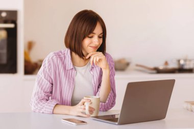 Remote job oppurtunities. Excited lady working on laptop and drinking morning coffee, sitting at table in kitchen interior, free space. Young woman having breakfast and using pc computer