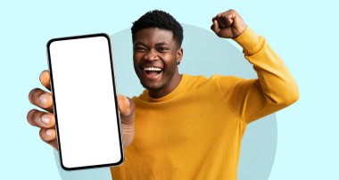 Parlak sarı bir süveter giyen genç bir adam kutlama için gülümsedi ve önünde açık mavi bir fona karşı akıllı telefonu tutarken yumruğunu kaldırdı. Onun sevinci başarıyı yansıtır.