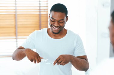 Yakışıklı siyah adam diş fırçasını diş fırçasına sürerken banyoda aynanın yanında duruyor, mutlu Afrika kökenli Amerikalı erkek dişlerini fırçalıyor, sabah temizliğini evde yapıyor, seçici bir odak noktası oluşturuyor.