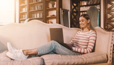 Oturma odasındaki kanepede oturup dizüstü bilgisayar kullanan neşeli bayanın portresi. İnternette arkadaşlarıyla sohbet eden mutlu bir kadın, klavyeye yazı yazan, evden çalışan serbest çalışan bir kadın.