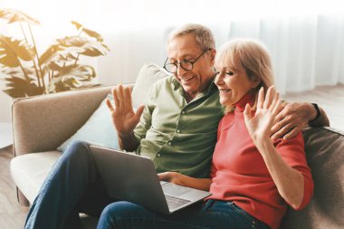 Mature Couple Waving Hands Video Calling Via Laptop Communicating Online Sitting On Couch In Living Room At Home. Spouses Gesturing Hello Talking To Computer Webcam. Distance Communication Concept