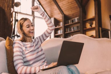 Büyük Galibiyet. Neşeli Pozitif Kadın 'ın Portresi Dizüstü bilgisayarla Sıkışan Yumruk Zevk ve Heyecan İçin, Elini Kaldırıyor. Evdeki koltukta oturan mutlu genç bayan. Evet, harika haberler ve zafer konsepti.