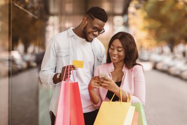 Kolay Ödeme Konsepti. Mutlu Afro-Amerikan erkek ve kadın portresi cep telefonunu birlikte kullanıyor. Kredi kartı ve alışveriş torbaları tutuyor. Alışveriş merkezinin yanında sıradan bir çift bekliyor. Perakende ve Satın Al