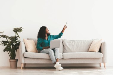 Şık giyinmiş mutlu siyah kadın evinde dizüstü bilgisayarla koltukta oturuyor, uzaktan kumandayı tutuyor, klimayı açıyor, internette sörf yapıyor, beyaz duvar arka planında, kopyalama alanının fotokopisini çekiyor.