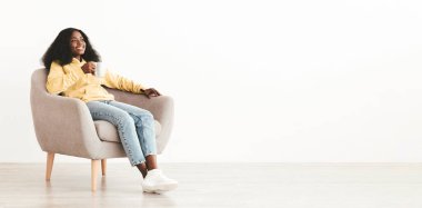 Rahat, bin yıllık siyah bir kadın rahat, gür saçlı, rahat koltukta oturuyor, beyaz duvarın üzerinde bitki çayı içiyor, fotokopi alanına bakıyor ve gülümsüyor, panorama