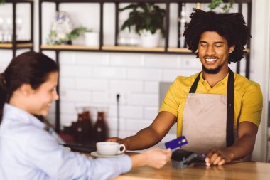 Bin yıllık Afro-Amerikan Kıvırcık Erkek Garson müşterisine taze kahve veriyor. Tatmin olmuş kadın da kafenin içindeki barın girişinde terminal ve kredi kartıyla ödeme yapıyor. İyi hizmet, iş.