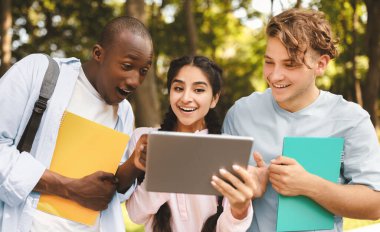 Kampüs eğlencesi. Parkta yürüyen mutlu uluslararası gençler, tabletini paylaşan Hintli bayan, arkadaşlarına yeni eğitim uygulamalarını gösteriyor, birlikte cihaza bakıyor ve gülümsüyor.