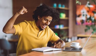 Mutlu, mutlu, heyecanlı, binlerce yıllık Afro-Amerikan kıvırcık adamı zafere seviniyor ve masada bilgisayar ve kafenin içindeki not defteriyle harika sonuçlar elde ediyor. Başarı ve iyi anlaşma, jest ve kazanmak