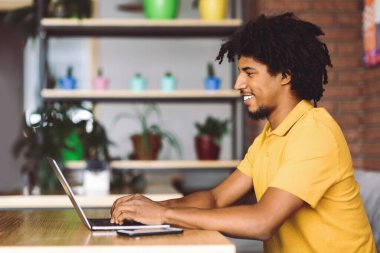 Kafenin içinde akıllı telefon ve profil ile bilgisayar klavyesinde yazan mutlu, kıvırcık, genç, siyah bir erkek. Uzaktan iş, iş için modern teknoloji, bağımsız müteahhitler için işyeri