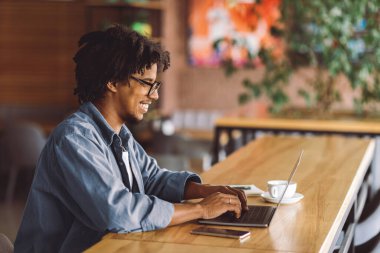 Neşeli gülümseyen kıvırcık milenyum gözlüklü siyah adam, kafenin içindeki masada dizüstü bilgisayarda yazıyor, kopyalama alanı. Dijital göçebe, serbest çalışma, birlikte çalışma ve öğrenim yeri, modern teknoloji