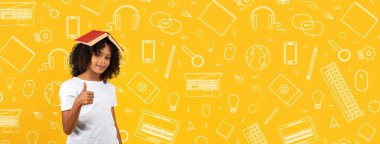 Neşeli bir çocuk kafasında bir kitapla poz veriyor, gülümsüyor ve başparmağını kaldırıyor. Parlak sarı arka planda okulla ilgili ikonlar var, enerjik bir atmosfer yaratıyorlar..